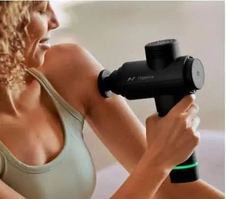 Woman using a black massage gun on her shoulder against a neutral background
