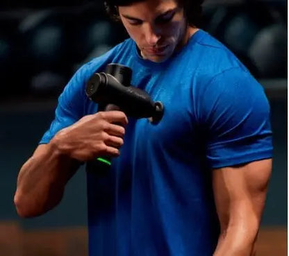 Man in blue shirt using a black massage gun on his arm against a dark background
