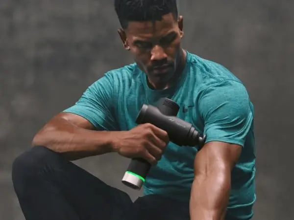 Man in teal shirt using a massage gun against a gray background