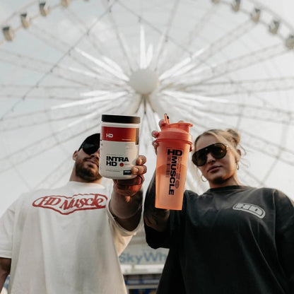 Two people holding HD Muscle supplement containers with a ferris wheel in the background