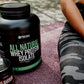Protein supplement bottle labeled 'All Natural Whey Protein Isolate' on a surface with a person wearing camouflage pants in the background.