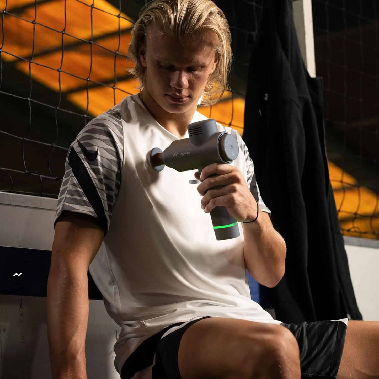 Erling Haaland using a Hyperice massage gun in a sports setting with a net in the background