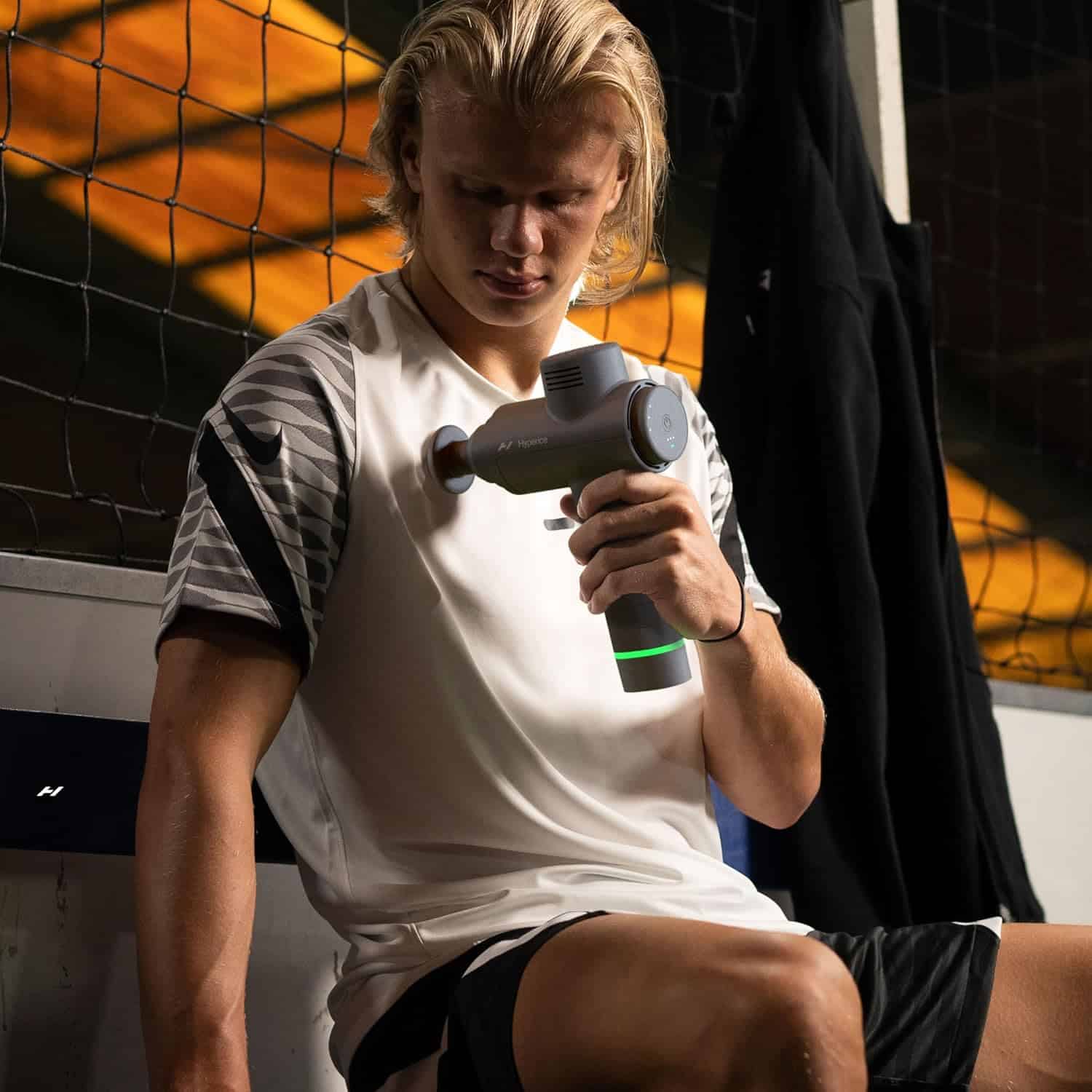 Erling Haaland using a Hyperice massage gun in a sports setting with a net in the background