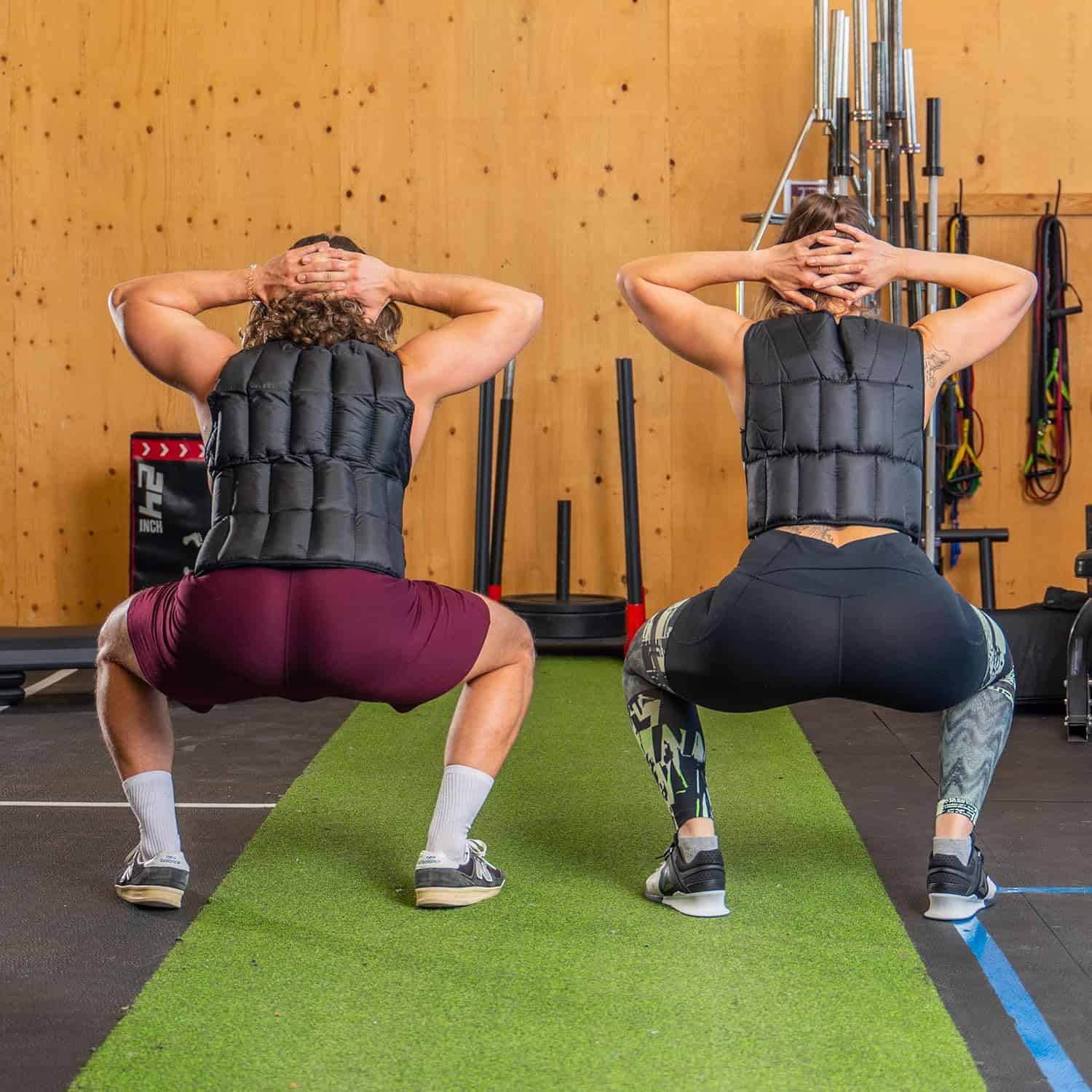 Two people performing squats with weighted vests in a gym setting.