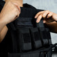 Person adjusting a black tactical vest with multiple pouches on a gray background